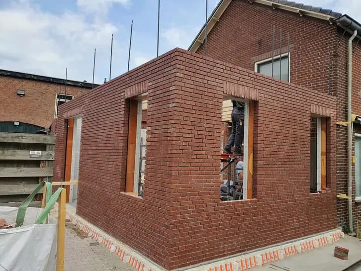Aanbouw van een woning in rood metselwerk tijdens de ruwbouw, met open sparingen voor kozijnen